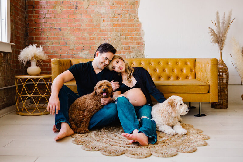 Personalized family portraits in Albuquerque with this joyful image of a small family expecting their first baby. They are seated in their living room in front of a brown couch, accompanied by their two beautiful dogs.