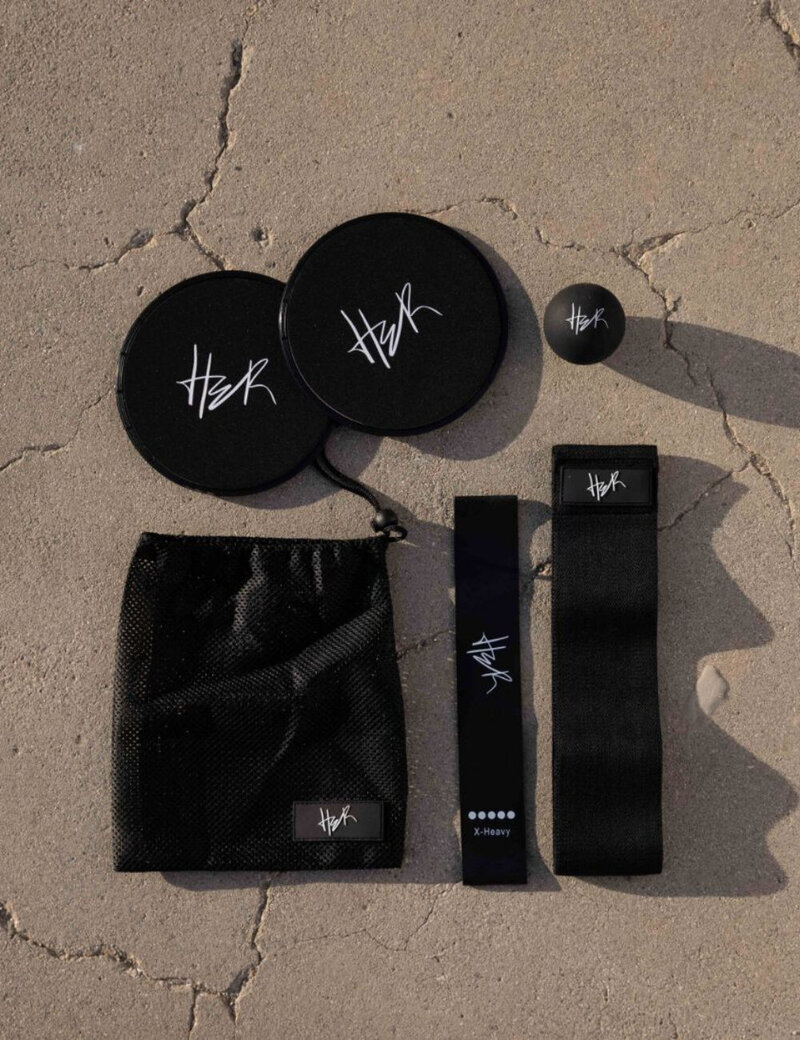 Flat lay of the HER Tool Kit including resistance bands, sliders, lacrosse ball, and carrying pouch on a concrete surface.