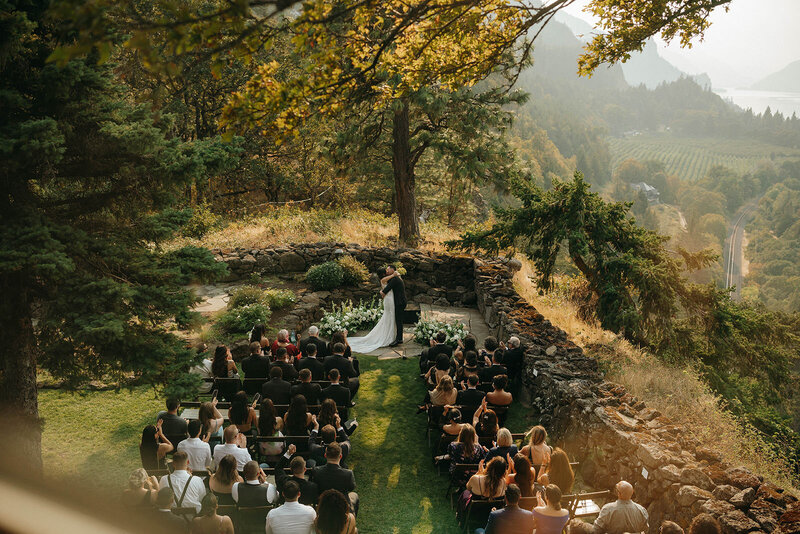 Outdoor wedding ceremony on a mountaintop with guests seated on a grassy terrace, surrounded by trees and overlooking a lush valley and river in the distance.