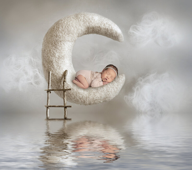 newborn baby girl wearing soft pink outfit sleeping in felt moon with reflection in the water