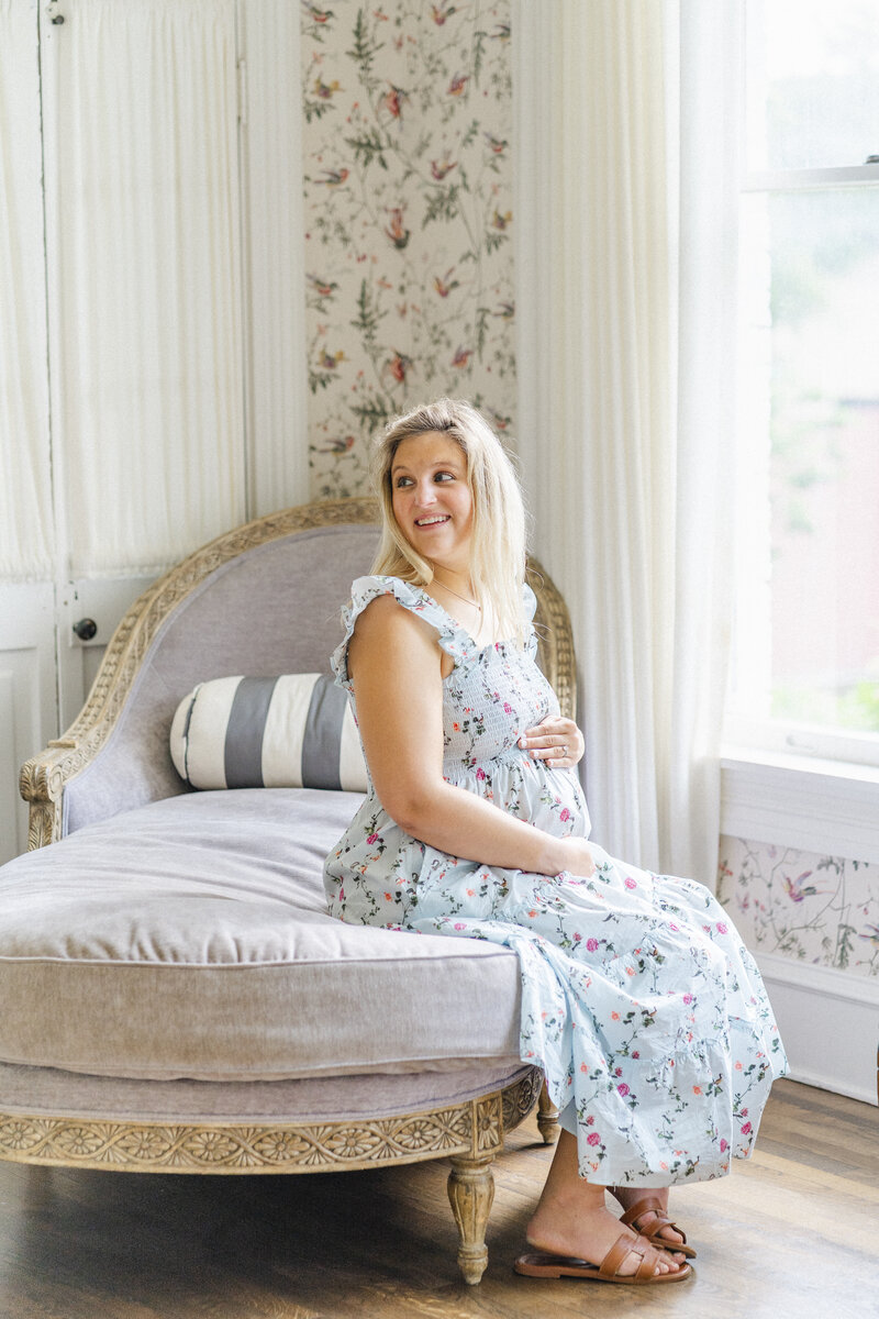 A pregnant mother in a floral dress sits on a daybed at Woodbine Mansion in Round Rock