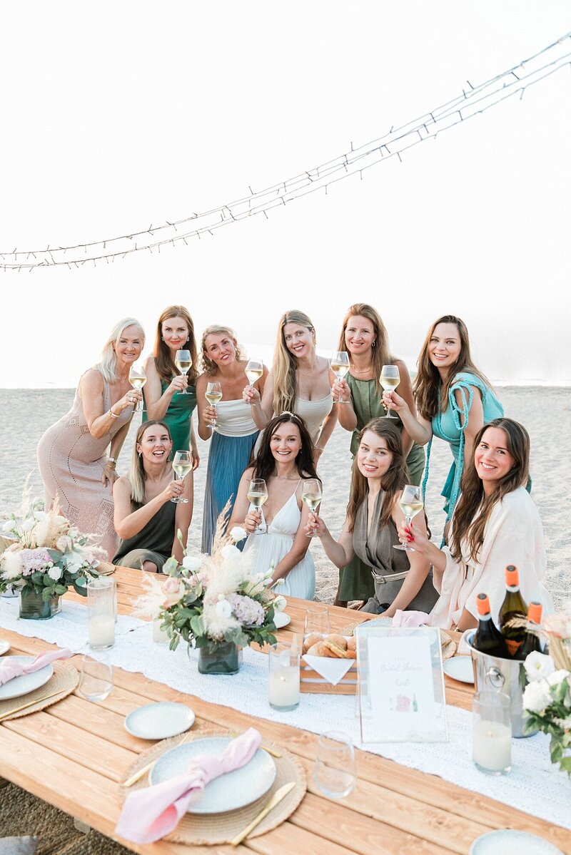 Bride celebrating with her bridesmaids and guests at a beachside welcome dinner, toasting with wine around a floral-decorated wooden table at sunset