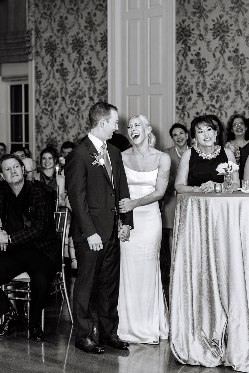 Black and white flash photo of bride and groom smiling at each other with guests behind them