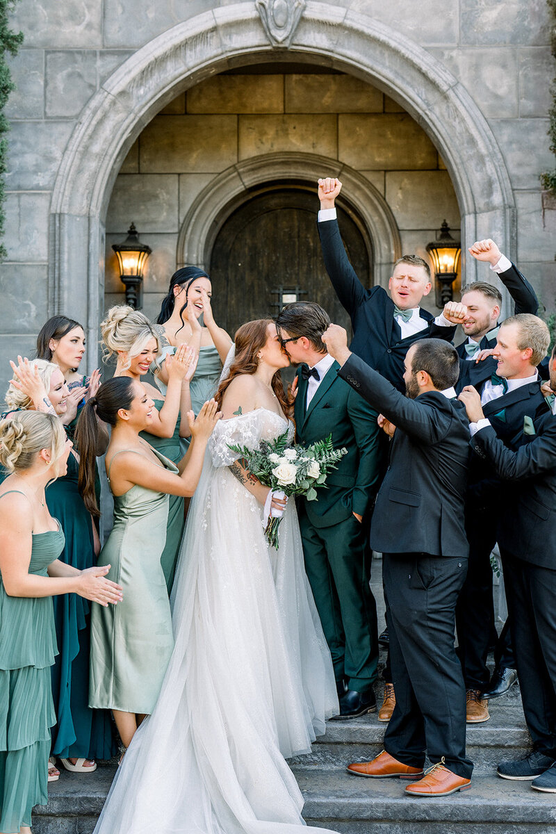 A joyful California wedding captured in a candid moment of celebration and love. The newlyweds share a kiss while surrounded by their cheering wedding party in shades of sage and emerald. This California wedding showcases a mix of modern elegance and timeless tradition, with heartfelt moments and an effortlessly chic bridal look. The outdoor setting provides a romantic backdrop, perfectly reflecting the warm, vibrant spirit of the occasion.