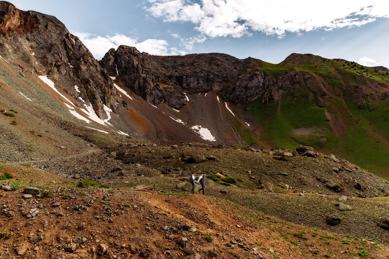 Shannon Lee, a Colorado elopement photographer capturing a couple walking across a rugged mountain landscape with towering peaks and snow patches in the background.