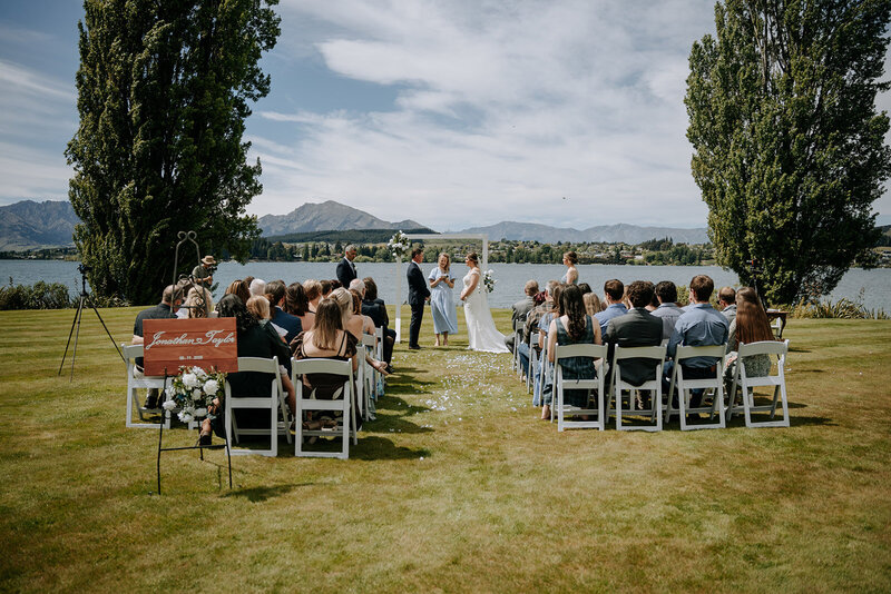 Bride and groom exchanging rings during a joyful lakefront wedding ceremony, officiated by a local celebrant with mountains in the background.