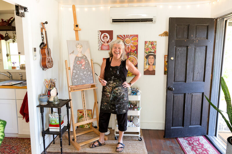Calgary brand storytelling photoshoot of a smiling female artist with silver hair standing in her cozy, well-lit studio wearing a black paint-stained apron. She holds a paintbrush in one hand, with a large easel displaying a whimsical portrait painting beside her. The walls are adorned with colourful, detailed artwork, and shelves and tables are filled with brushes, paints, and creative supplies. The open black door lets in natural light, adding warmth to the inviting, artistic space.