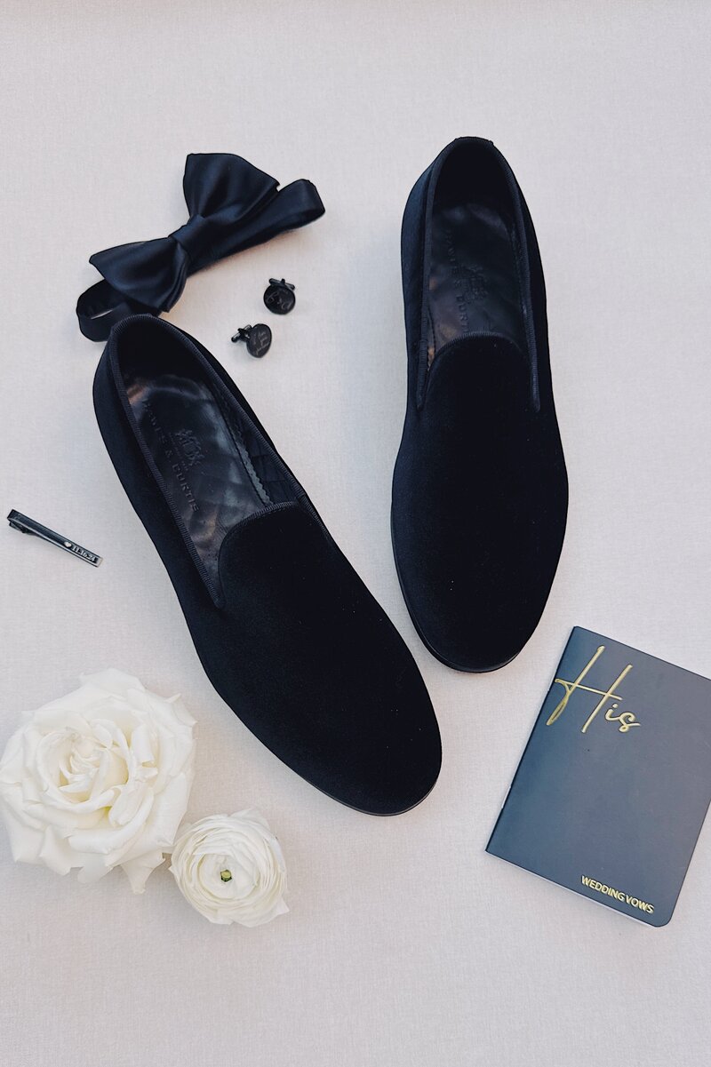 Groom details including black suede shoes, black vow book, white roses, bow tie, cuff links, and tie clip on the balcony of La Valencia Hotel.