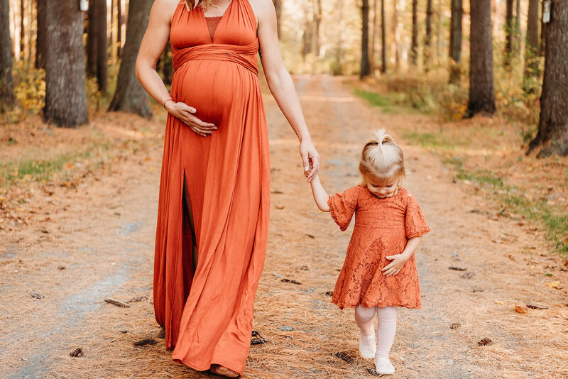 pregnant mom holding her young daughter's hand and walking towards the camera.  Mom is using her other hand to cup the under side of her baby bump, while daughter does the same, trying to imitate mom.  photo taken during Delaware maternity photography session.