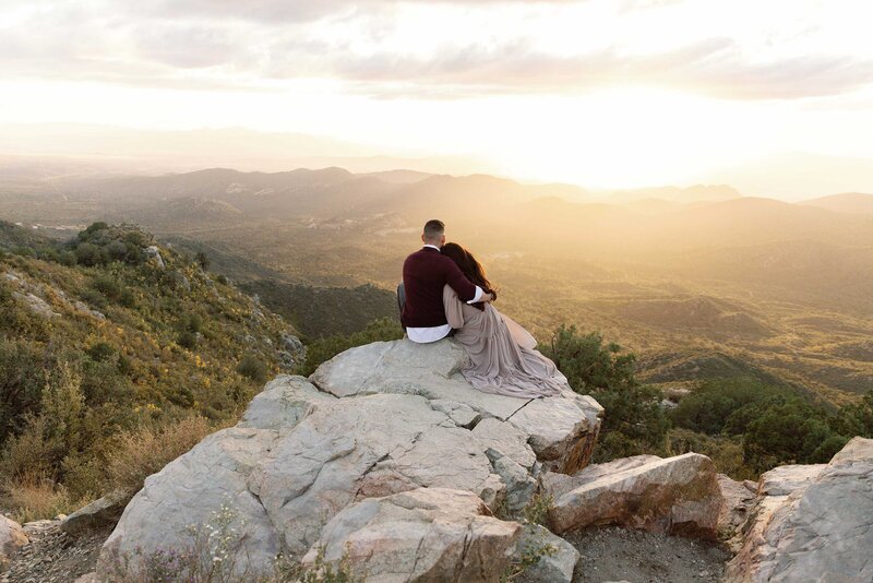 Couple sits cuddled together on cliffside.