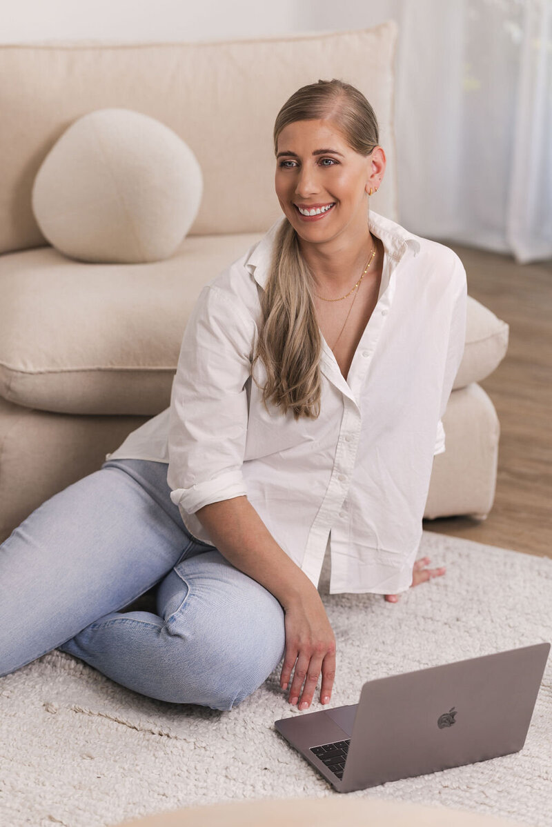 Woman smiling while wearing a white shirt and jeans sitting on a rug with a laptop.