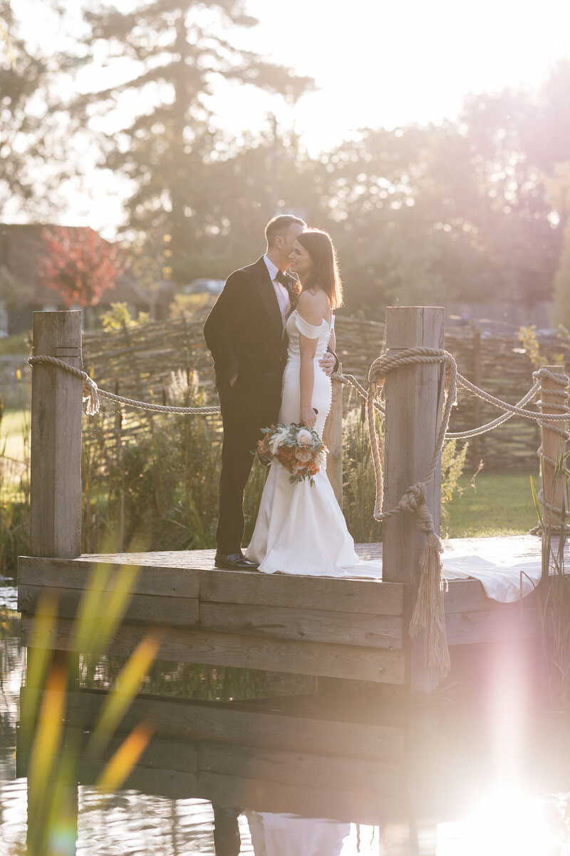 A bride and groom stand on a wooden dock by the pond at Silchester Farm, embraced by lush greenery as the sun sets. The bride holds a bouquet in her flowing white dress, while the groom in a dark suit tenderly kisses her forehead. Sunlight filters through the trees, casting a warm glow.