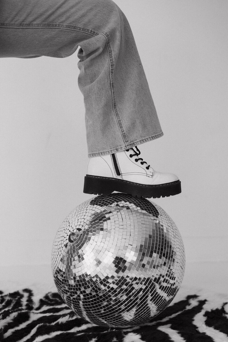 White combat boot stepping on a mirror ball placed on top of a zebra carpet in front of a white background