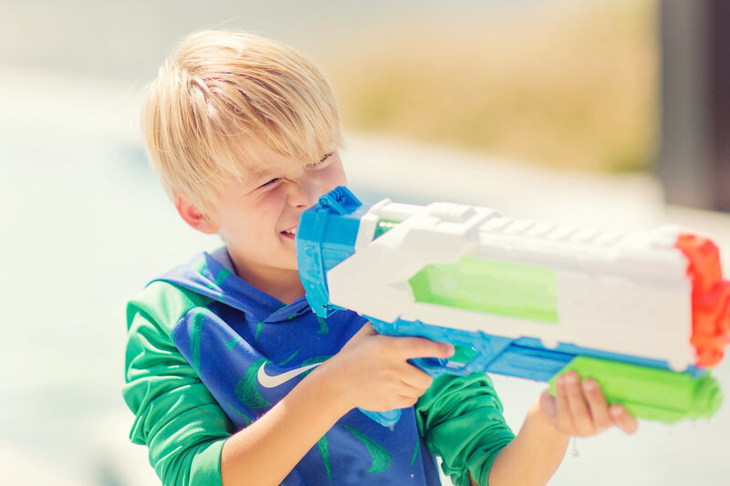 Young boy is playing with a big squirt gun in his backyard, aiming at his target.