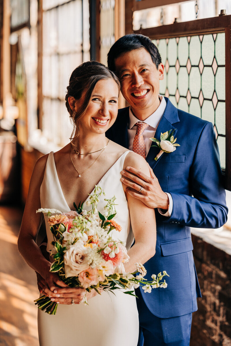 Brunette bride with spring floral bouquet smiles with groom wearing blue tux.
