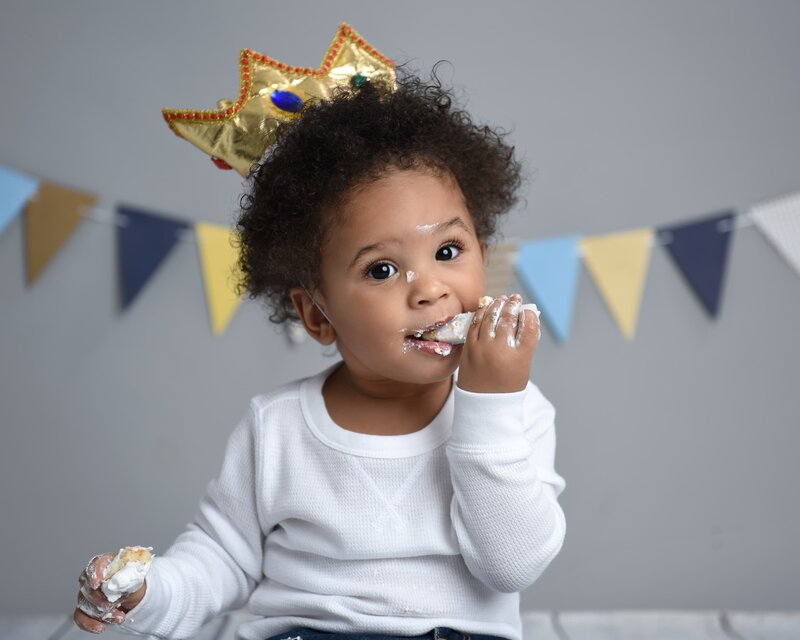 A baby boy wearing a gold crown and white outfit, enjoying his cake with frosting on his face, in a royal-themed setup with bunting in the background.