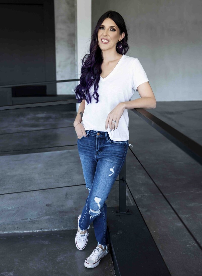 Laura stands, leaning into the corner of a black railing behind her. She smiles at the camera, with her right hand in her jeans pocket, and her left arm casually resting on the railing. She is wearing a white v-neck t-shirt, distressed jeans, and white sneakers. Her long hair is curly and purple with dark brown roots, slung over her right shoulder.
