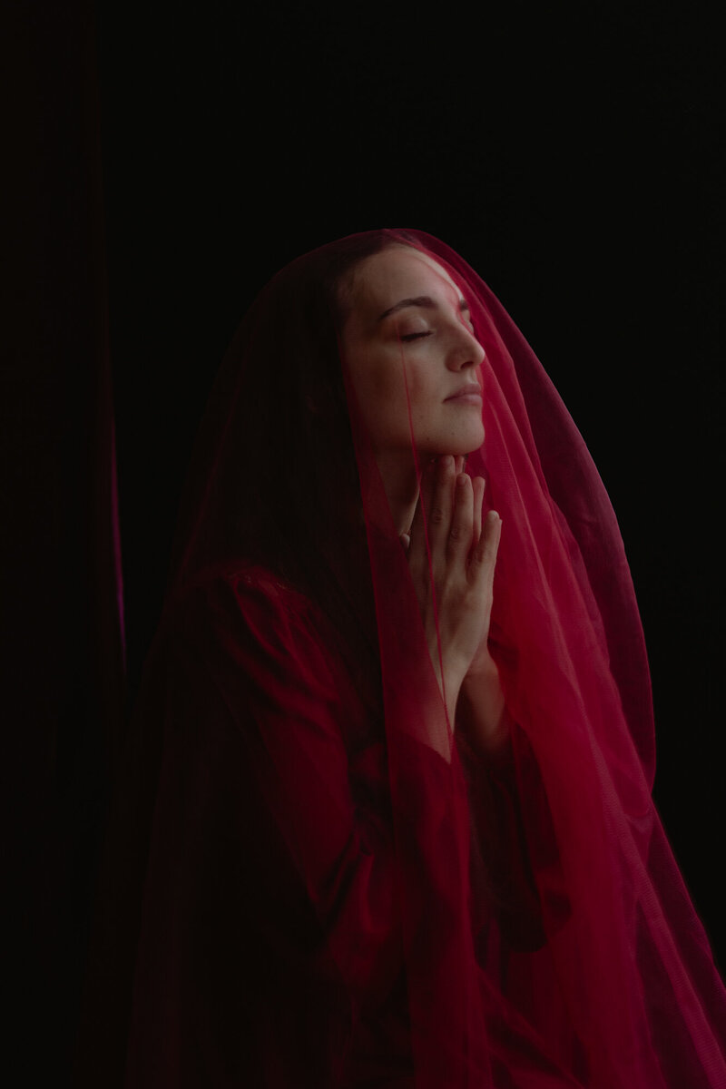 Donna seduta vicino a una finestra con luce soffusa, occhi chiusi e mani sul cuore, immagine di calma e centratura.