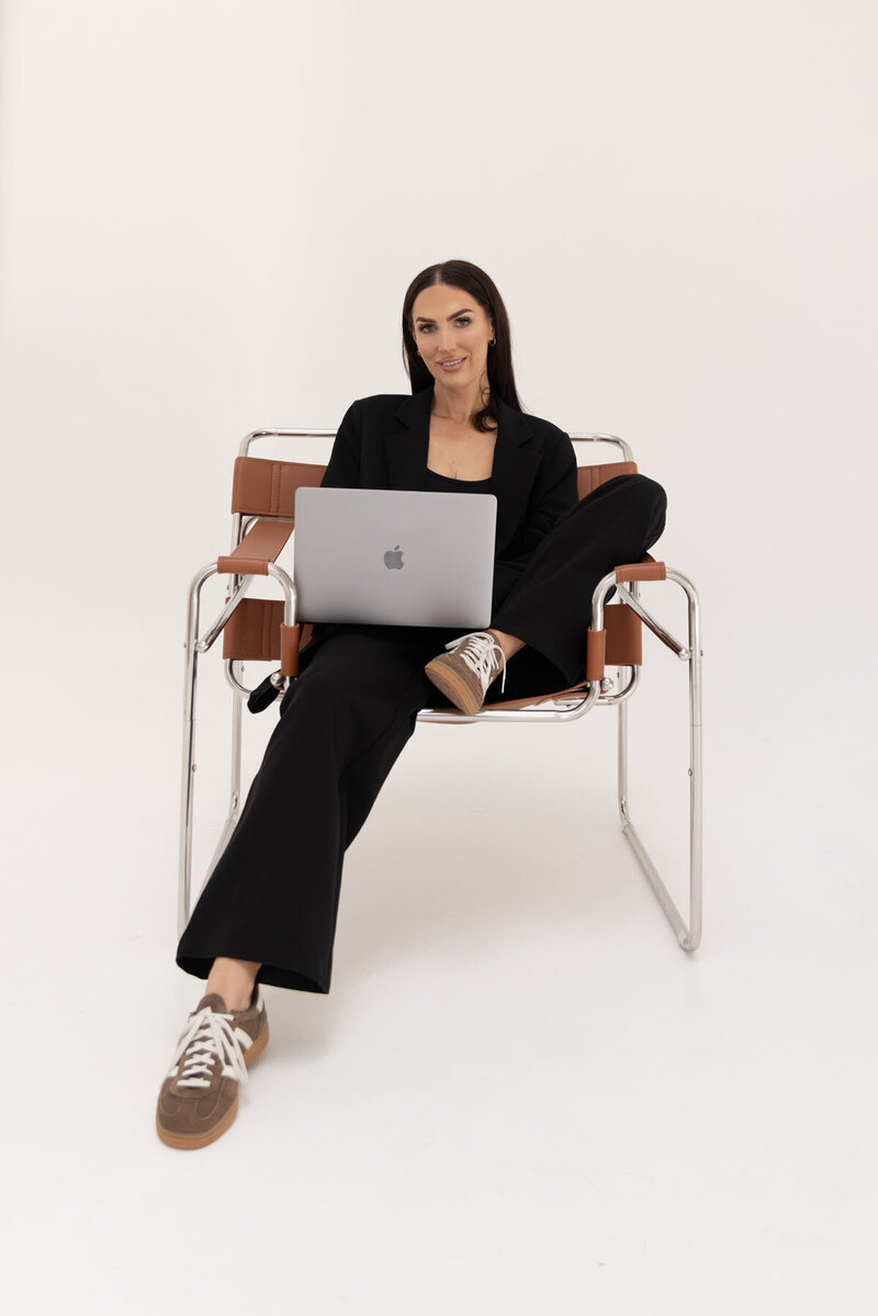 Tayla from Aurum Design Studio sitting casually in a chair with a laptop, wearing a black blazer