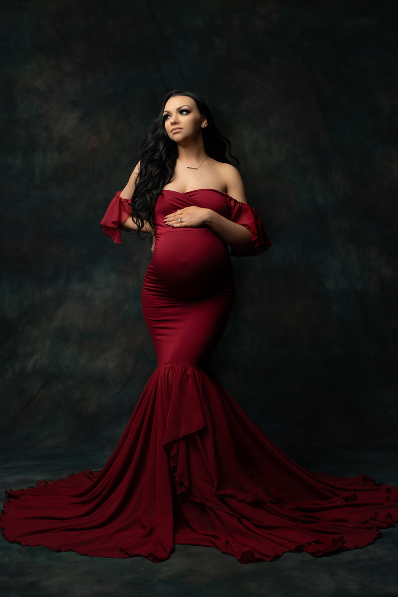 High end maternity portrait on dark grey backdrop with pregnant woman wearing burgundy form fitting dress that is flowy at the bottom