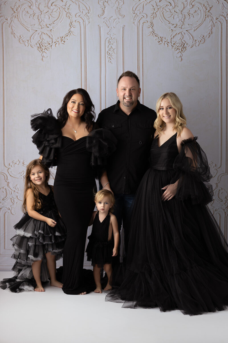 A family of five poses in an elegant studio wearing black formal attire with tulle gowns and dramatic sleeves. The parents and three children are smiling against a soft ornate backdrop. Captured by a Dallas Family Photographer.