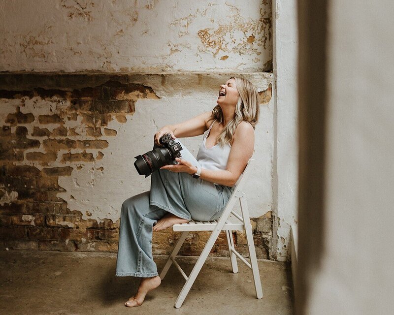 Image of Perth Family and Newborn Photographer, Kaz Soom.  She is wearing faded jeans and a white top, sitting in a white folding chair holding her camera and throwing her head back in laughter. 