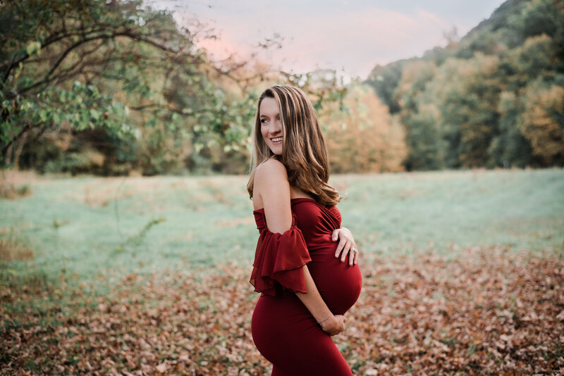pregnant blonde woman wearing wine red dress holding baby bump and smiling and looking back with fall setting leaves on ground