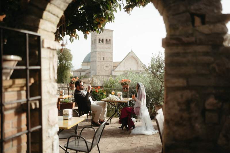 Framed by a stone archway, groom takes a photo of the bride with a basilica behind them for a fun Elopement Photo Idea
