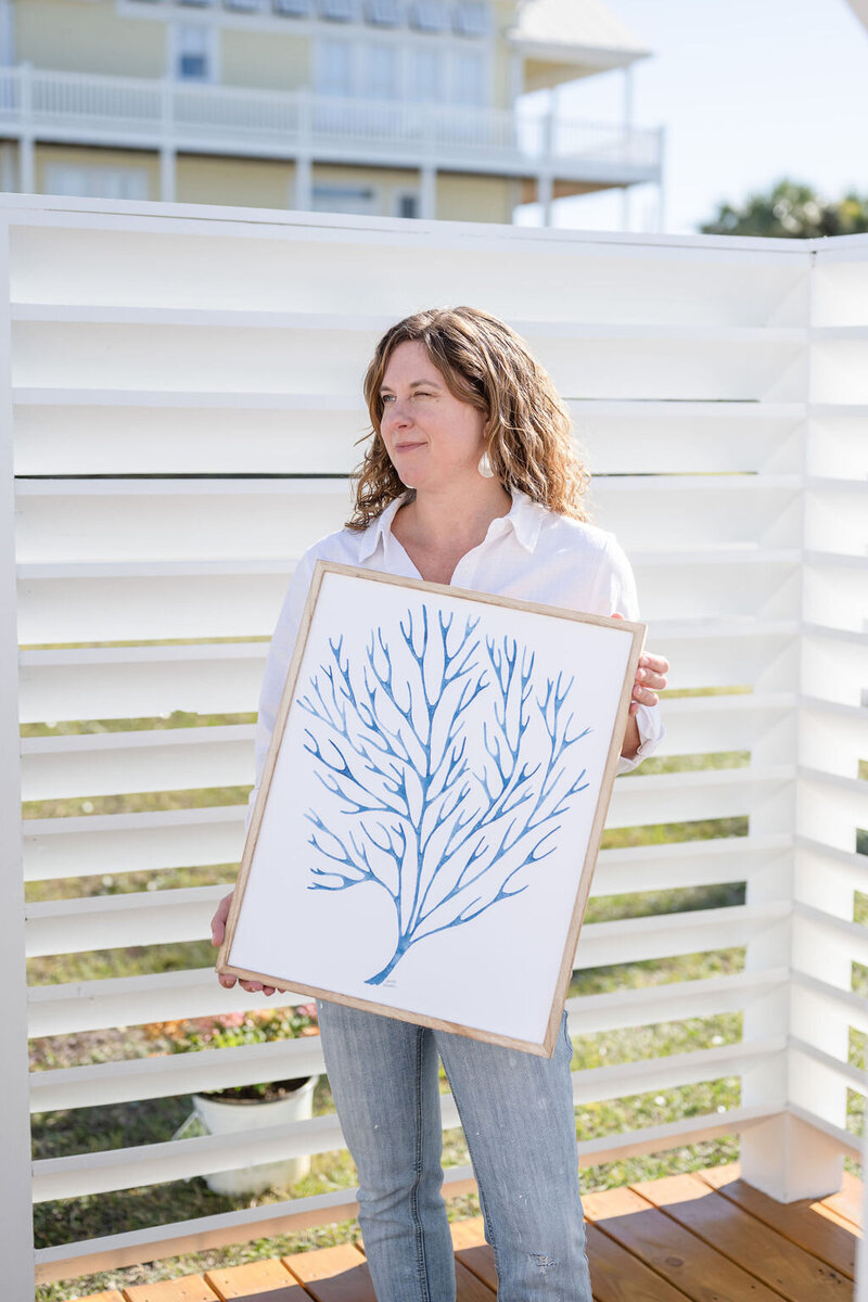 Laura, a watercolor artist holding a framed canvas of her blue marine plant painting in the sunlight