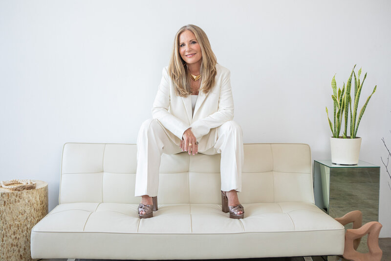 woman in a white suit sitting confidently on the back of a cream-colored couch in a modern indoor setting