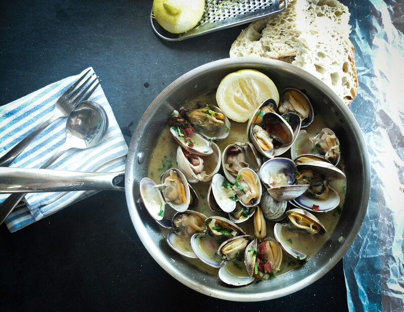 A pan filled with cooked clams in a broth, garnished with herbs, half lemon and slices of bread are placed beside the pan, blue and white striped napkin with a spoon and fork rests on the left