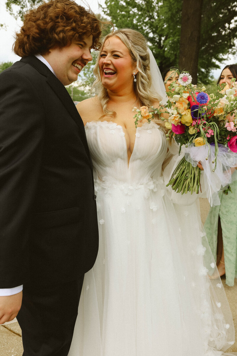 bride and groom hold ands among wildflowers in grassy field with green summer trees in background during elopement in Columbia, MO, wedding.