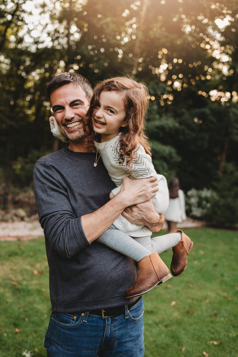 A joyful father holds his laughing daughter in an outdoor setting during a warm and candid family photography session in Connecticut. Captured by Nicole Mele Photography, highlighting love, connection, and genuine moments.