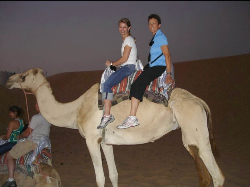 Two people are sitting on a camel in a desert setting during dusk. They are smiling and appear to be enjoying the ride. Another camel with riders is visible in the background. The sky is a dim, dusky color.
