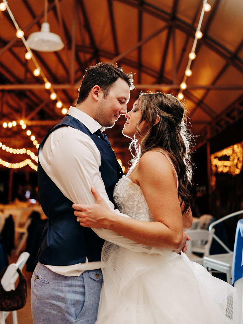 A bride and groom, captured by a wedding photographer in Eau Claire, embrace and smile lovingly at each other under warm string lights at an indoor wedding reception. The bride wears a white dress, and the groom is in a vest and tie.



