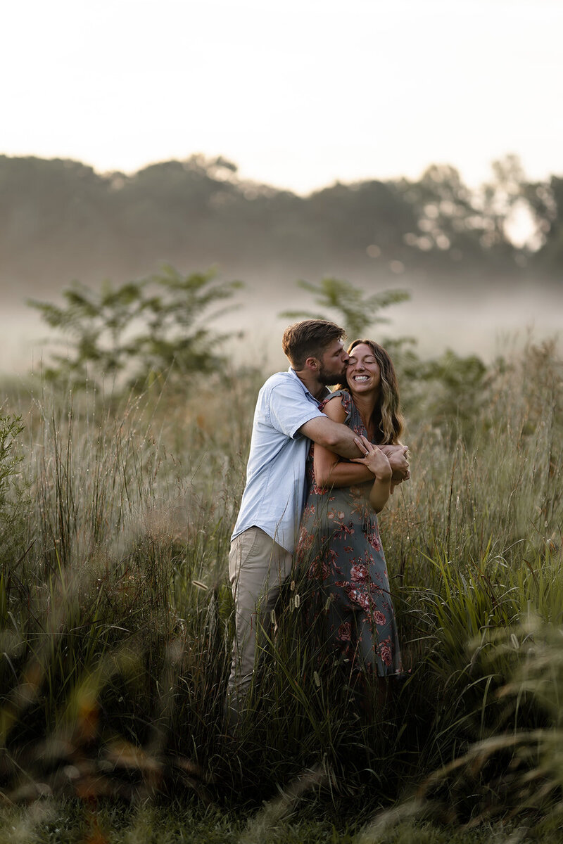 A couple cozied up standing in a field of tall grass in the early morning with fog in the background and dew on the grass 