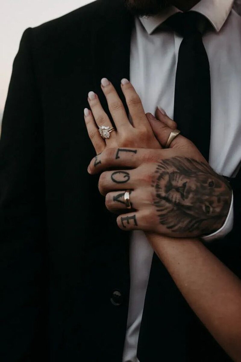Close-up photo of a bride and groom's hands intertwined. The bride's hand, adorned with a diamond ring, rests on top of the groom's hand, which features a tattoo of a lion and the word "LOVE" across his knuckles. The groom is wearing a dark suit and tie.