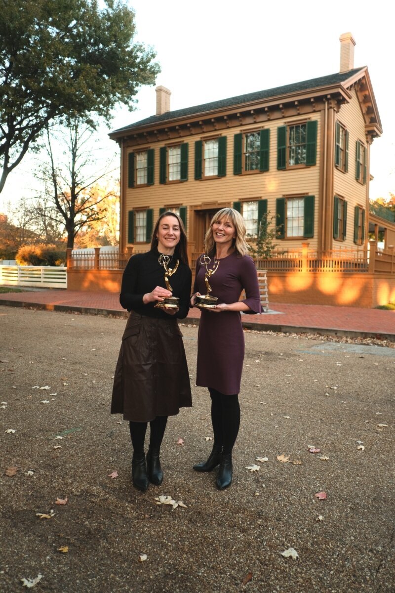 Two women holding Emmy® Awards stand smiling in front of the historic Lincoln Home in Springfield, Illinois, during golden hour.