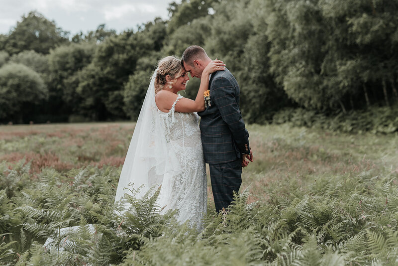 Couple gently stand forehead to forehead amongst the fern leaves in the meadow at Two Woods Estate, Pulborough