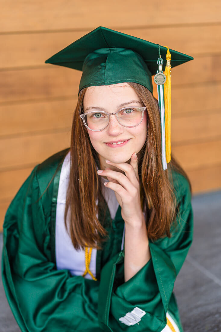 A closeup of a high school senior girl wearing a royal green colored graduation cap and gown sitting in front of a wooden wall in downtown Provo