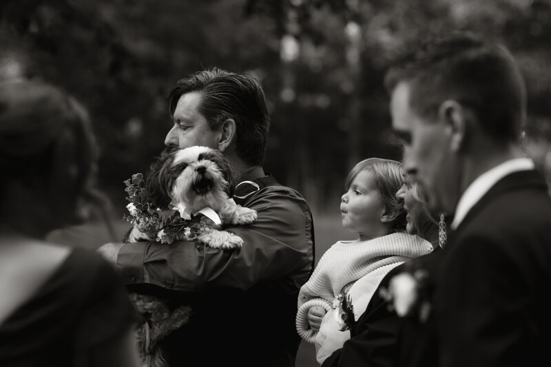 Black and white image of a man holding a small dog wearing flowers beside a child during a wedding ceremony, captured with emotional depth by Rainstorm Photo & Video.