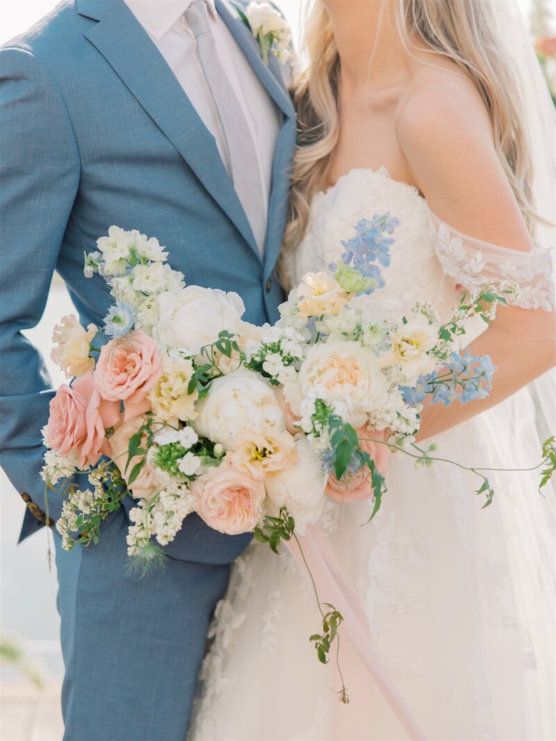 Close up of a bride and groom, shoulders to knees, showing off the bride's bouquet at they lean against eachother