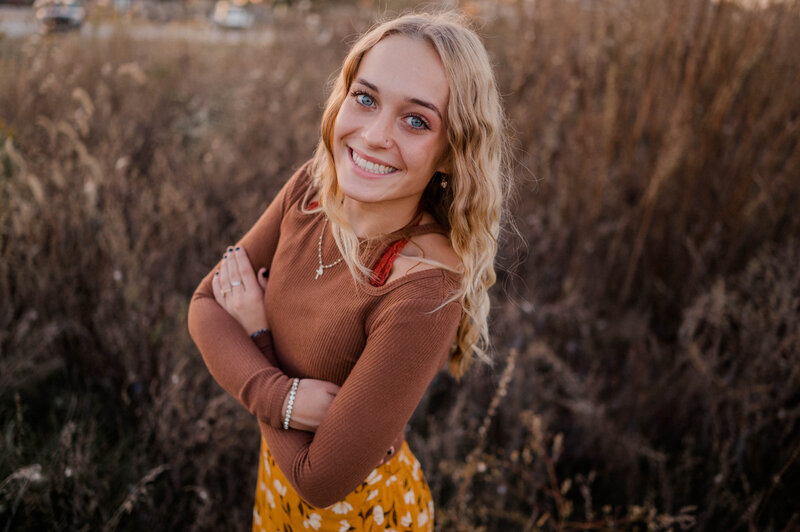 A high school senior girl poses with her arms crossed and smiling at the camera while standing in an open field.