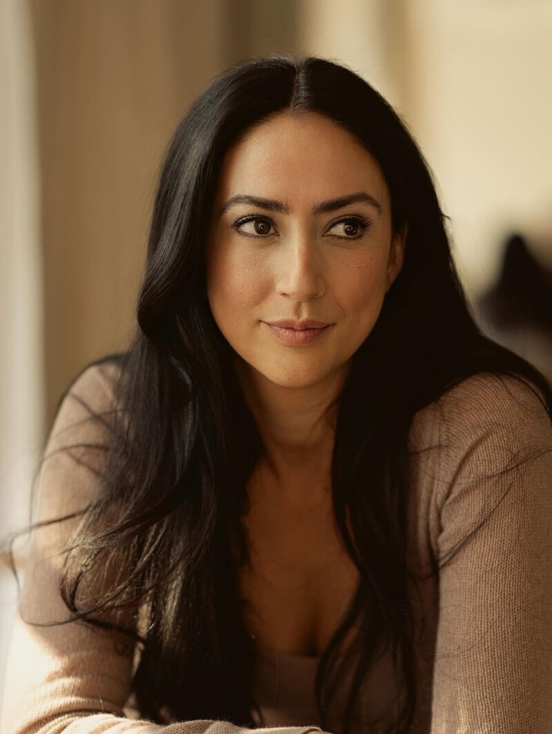 A woman with long dark hair leans on a white chair, resting her chin on her hand and gazing thoughtfully to the side. She wears a brown sweater and sits in a softly lit, neutral-toned room.