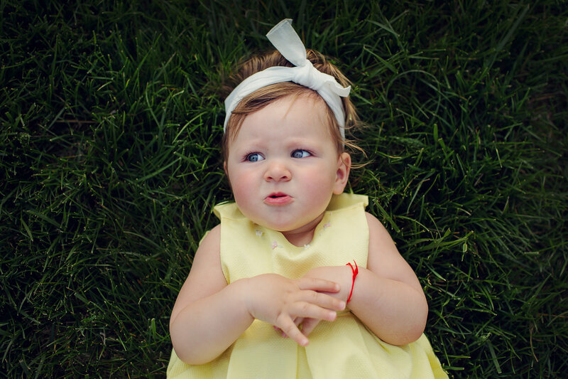 Baby girl is laying in the grass in a yellow dress and white bow in her hair with a sad face.
