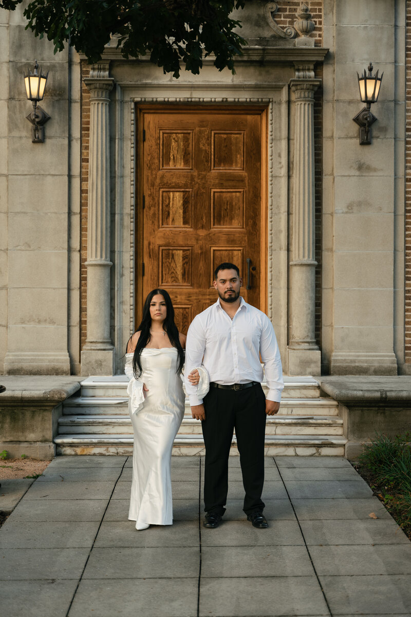 A stylish couple in sunglasses stands in front of an ornate, large wooden door framed by tall columns. The mood is elegant and sophisticated.