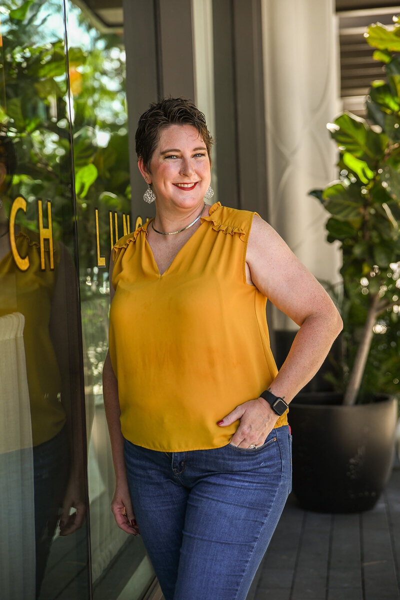 woman wearing a mustard yellow tank top and jeans poses in front of a cafe window.