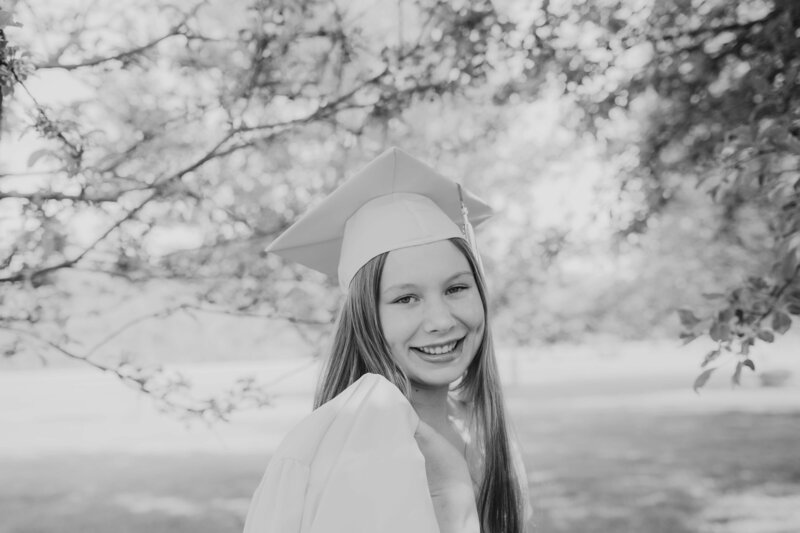 Black and white senior photo of a smiling high school graduate wearing a cap and gown in an outdoor setting, captured during a Michigan Ohio senior photography session celebrating graduation season.