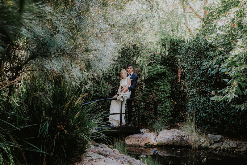 Bells at Killcare is the best Boutique Luxury Elopement Venue on the Central Coast. The image shows a married couple on their wedding day perched on a small bridge over a pond surrounded by trees. It is elegant and has a natural Australian feel.