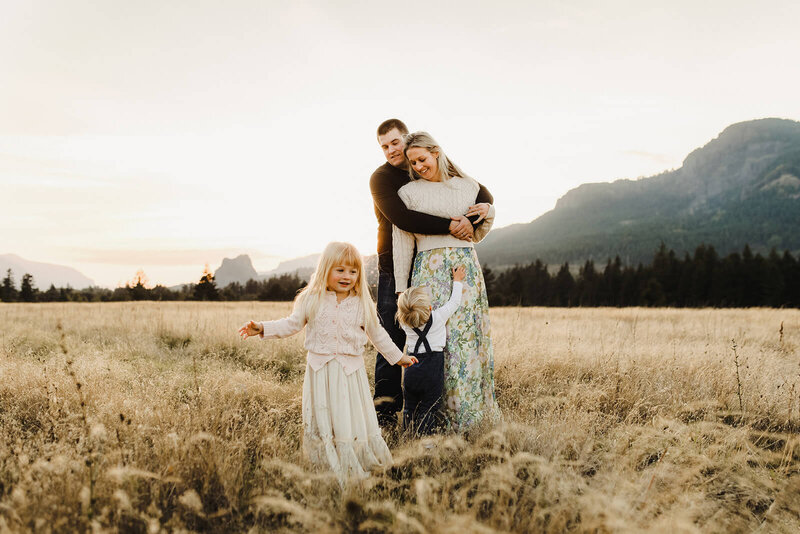 A dad wraps his arms around his wife while they smile down lovingly at a toddler at their feet, in the foreground a little girl explores the field they are standing in in the Columbia River gorge